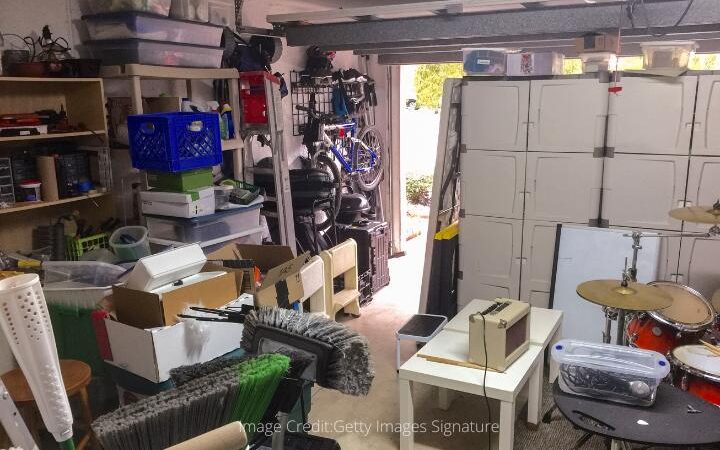 messy garage interior with brooms, a beige amp on a white table, a red drumset in front of beige containers that are stacked up 5 feet high, shelves of cables, plastic containers, and shoe boxes, a bike on a rack