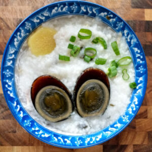 image: bowl of black, preserved duck egg cut in half over rice porridge, slice of ginger, and sliced green onions in a blue bowl on a wood cutting board